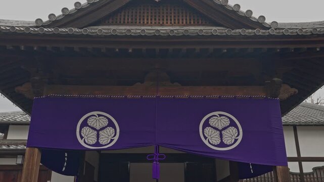 ilt-down shot revealing a vivid purple ceremonial banner with twin white family crest motifs beneath a traditional dark wooden shrine hall roof, overcast grey sky above.