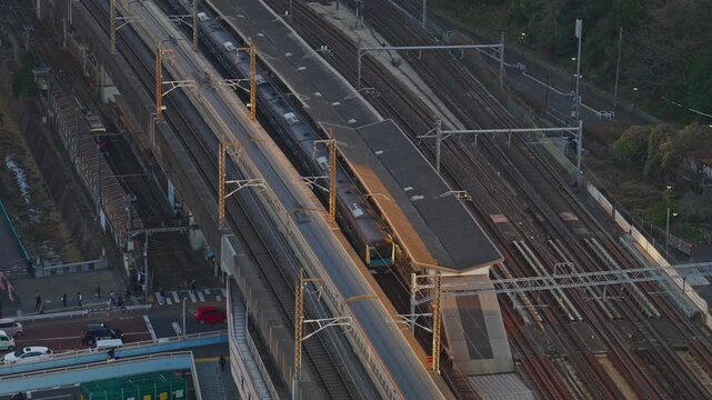 High-angle static aerial shot of a blue and white high-speed train on parallel tracks in a busy railway yard, overhead catenary wires, warm dusk sidelight across the rooftops below.