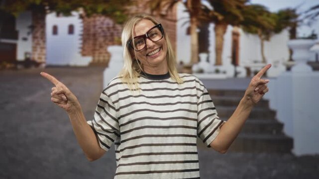 Woman, middle age blonde wearing large glasses and striped shirt, pointing both index fingers while smiling and showing hands at white building steps; cheerful playfulness.