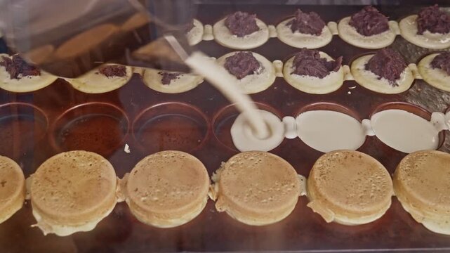 Fresh batter pours into oval iron molds alongside rows of imagawayaki at different stages &mdash; empty molds, batter-filled, and red bean paste-topped &mdash; showing the full street food production sequence.