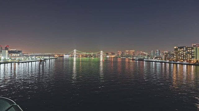 Ultra-wide static time-lapse of Tokyo Bay at night, Rainbow Bridge with green cables centered in distance, illuminated piers flanking both sides, small boat wake cutting through dark rippling water.
