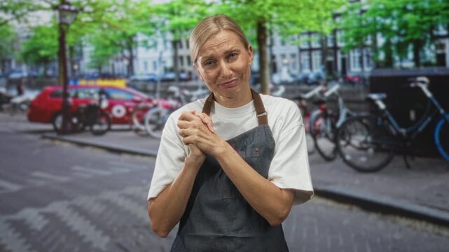Woman in apron clasping hands and smiling on street near bicycles and parked car; warmth hospitality approachability.