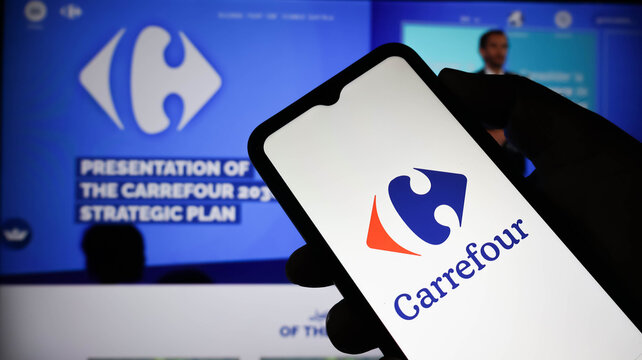 A person holding a smartphone displaying the carrefour logo in front of a presentation screen about the carrefour 2026 strategic plan