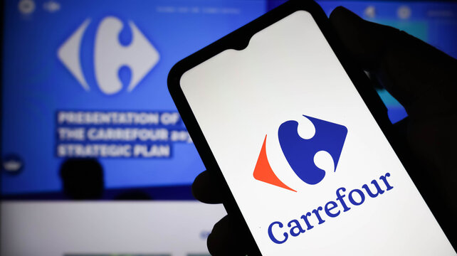 Closeup of a hand holding a smartphone displaying the carrefour logo on the screen with a presentation about the companys strategic plan in the blurred background
