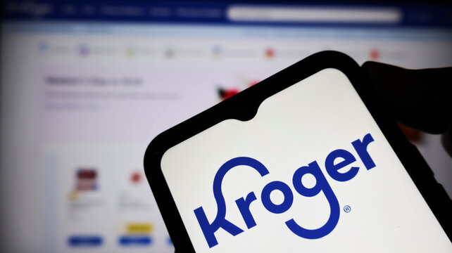 Closeup of a hand holding a smartphone displaying the kroger logo on the screen with a blurred computer monitor in the background
