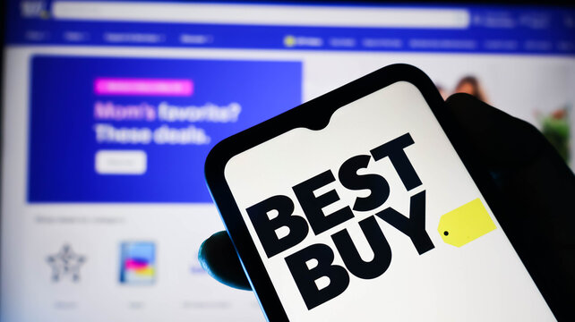 Closeup of a hand holding a smartphone displaying the best buy logo on the screen with a blurred computer monitor in the background