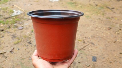 Naklejka premium Hand Holding Empty Brown Plastic Plant Pot on Outdoor Background, Gardening Container. Hand holding empty red plastic flower pot on ground background.