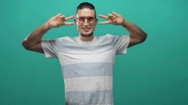 Man wearing striped t shirt and red glasses showing v sign with both hands by eyes in studio; playful youth.
