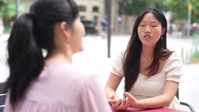 Two women sit at an outdoor cafe table. One smiles warmly, eyes focused on her companion. The other listens, head slightly tilted, showing attentiveness. Their hands rest gently on the table's surface