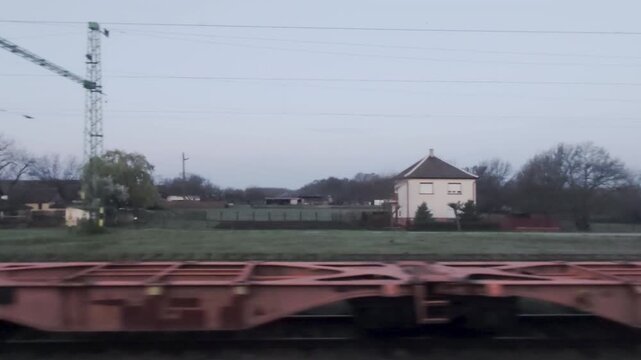 A professional tracking shot captures the industrial landscape of a Hungarian railway junction. Green catenary masts and a passing freight car contrast with a quiet village house under a pale morning