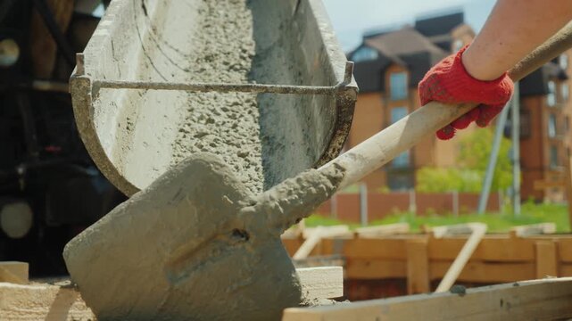 Worker using shovel to guide liquid concrete flow from mixer truck chute into foundation formwork during residential building construction on sunny day with motion of pouring materials at site.