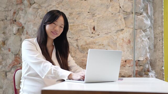 Young woman smiles while typing on laptop. She sits at table against rustic stone wall. Her focused gaze shifts from screen to camera. Her relaxed posture suggests calm productivity