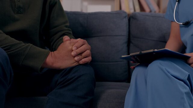 Compassionate nurse providing care, comfort to nervous elderly patient sitting on sofa during health conversation, holding clipboard, touching hand offering reassurance in homecare consultation