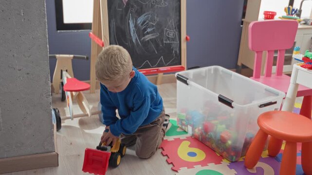Boy kneels holding red toy dump truck beside clear bin of blocks and chalk easel in building; quiet curiosity and childhood play.