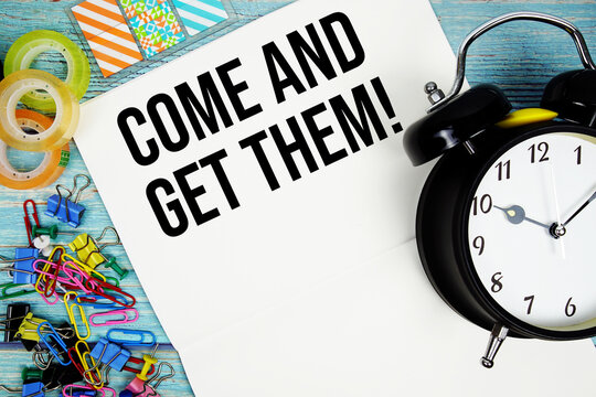 Come And Get Them! text with alarm clock and colorful office supplies on a blue wooden surface