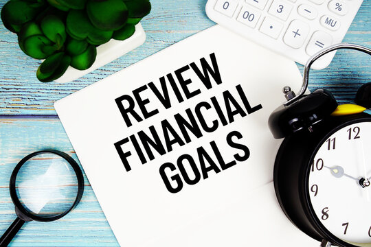 Review Financial Goals message written on paper placed on a blue wooden surface, alongside a magnifying glass, a clock, a calculator, and a small plant
