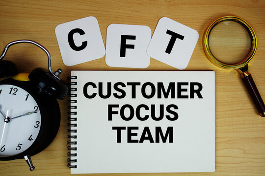 CFT Customer Focus Team text written on notepad with magnifying glass and alarm clock on wooden background