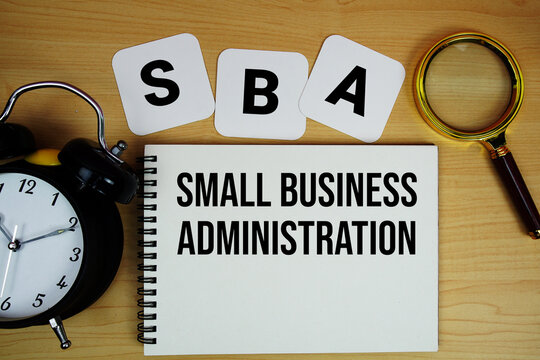 SBA Small Business Administration text written on notepad with magnifying glass and alarm clock on wooden background