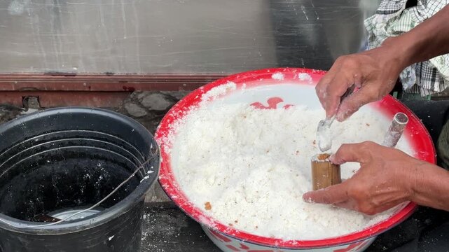 An Asian man selling traditional food fills a bamboo mold with Putu cake batter made from rice flour and palm sugar before the steaming process begins.