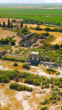 Vertical video. Balat, Turkey. Aerial view over the ancient ruins of Miletus, revealing the North Agora, Bouleuterion council house, and Nymphaeum amid sun-bleached stone and lush green fields. Aeria