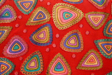 Fototapeta premium Close up of red fabric with colorful embroidered triangular patterns. Abstract textile background featuring vibrant geometric shapes and circular dot details.