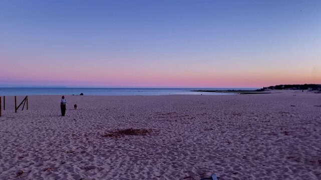Person walking dog along an expansive sandy beach during a vibrant sunset, showing tranquil coastal leisure and travel