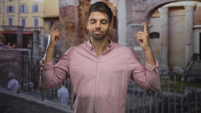 Hispanic man with beard pointing both index fingers up, bare hands visible, wearing pink button shirt before stone arch and metal railing on street steps; contemplation hopeful.