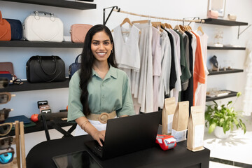 Indian boutique owner or worker stands behind checkout counter in clothing and accessories store, working on laptop, surrounded by handbags and garments n background. Retail services, entrepreneurship © fizkes