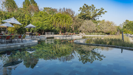 Obraz premium Outdoor swimming pool with rounded curved perimeter outlines. There are tables, chairs, umbrellas on the terrace. Pedestrian bridge over the water. Green tropical trees. Mirror reflection. Nepal. 