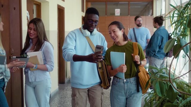 African male student and Korean female student smiling while looking at smartphone and walking through busy university hallway during class break.