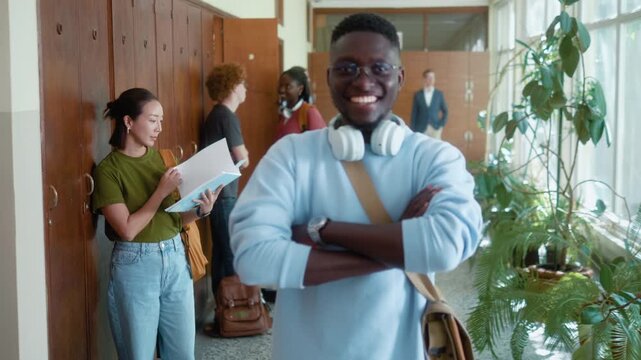 Smiling African male student with backpack looking at camera in university hallway during break with diverse classmates in background.