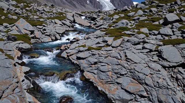 rocky alpine stream with glacier in background, turquoise rapids rushing through boulderstrewn valley under clear blue sky, towering snowcapped peaks and scattered rock slabs, moss patches clinging
