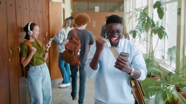Surprised African student celebrates passing exam after reading good news on smartphone in university hallway. Excited reaction, dancing and academic success.