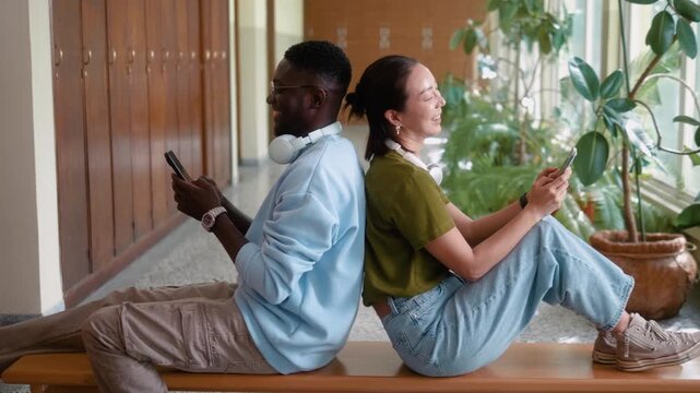 African male student and Korean female classmate sitting back to back using smartphones during break in university hallway. Modern campus lifestyle.