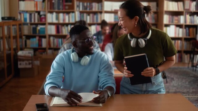Asian female student brings book and sits beside African classmate for collaborative studying in university library with diverse students in background.