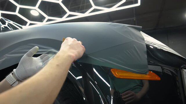 Close up view of a applying matte vinyl wrap on a car body panel in a workshop. Automotive detailing process with protective film installation, smooth matte finish and precision work.
