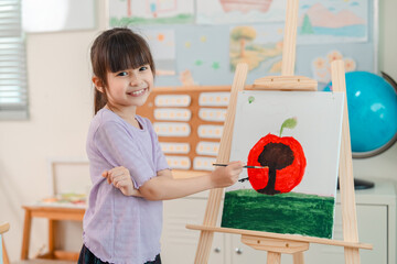 Obraz premium Back to school and education concept of smiling girl painting apple on easel in classroom art lesson