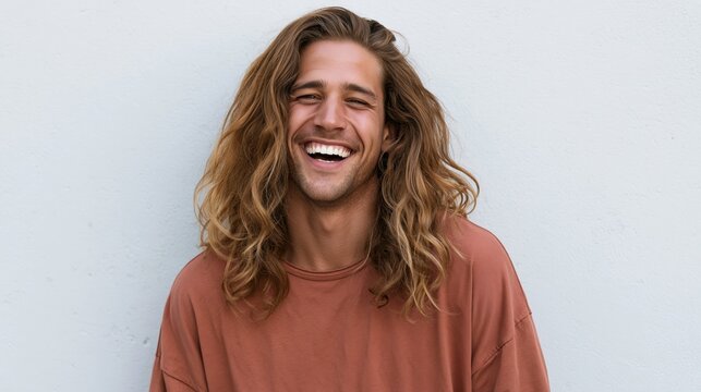 Handsome young man with long wavy hair laughing joyfully and showing a bright smile against a neutral background.