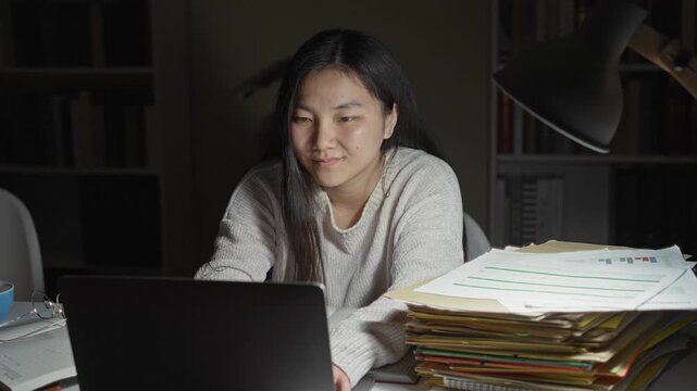 Young chinese woman resting chin on hand at laptop with stack of papers, cup and glasses, studying late at a desk inside a building; study break contentment.