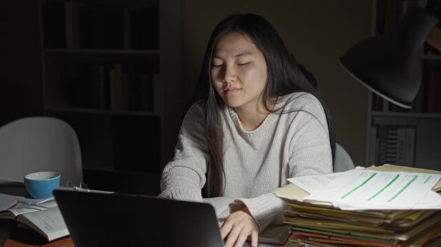 Woman studying at a desk with laptop, open notebook, stacked folders and mug, hands clasped in thought under a desk lamp in a building; late night fatigue.