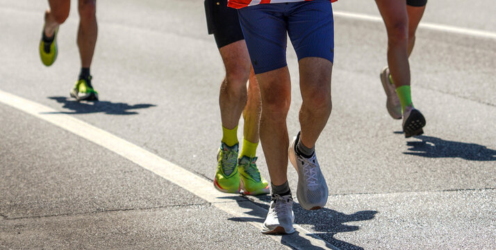 Road race runners approaching in bright sunlight with strong shadows on asphalt. Low crop of legs and running shoes shows marathon pace and endurance