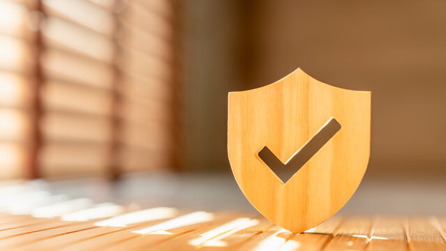 Quality assurance and protection concept. Wooden shield with checkmark icon on desk under sunlight. Ideal for internet security, data privacy, health insurance, warranty and certified compliance.