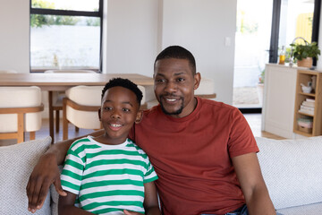 Fototapeta premium African American father and son sitting on upholstered couch in modern living-dining area smiling