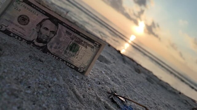 A video showcasing a dollar bill shape in the sand on a beach during sunset