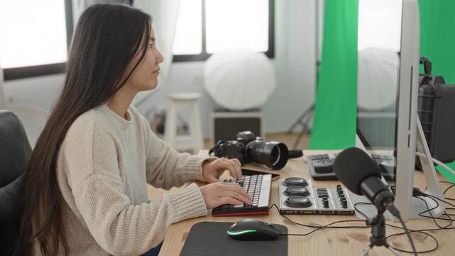 Woman photographer typing on keyboard beside camera and microphone in studio with green screen and long hair; concentration creativity.