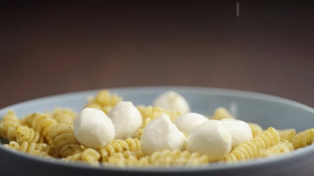 Slow motion mozzarella balls fall on pesto fusilli in blue bowl