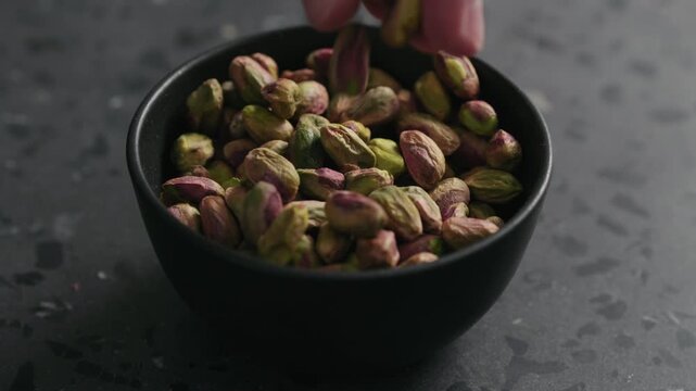Slow motion man hand takes pistachio kernels from black bowl on terrazzo surface