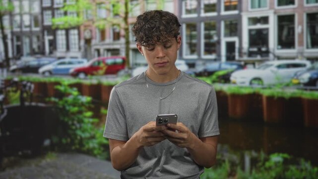 Teenage boy holding smartphone with fingers scrolling while looking down on a street canal by buildings; quiet thoughtful.