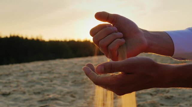 Hands pouring golden sand between fingers in slow motion at sunset close up shot of grains falling against warm backlit background creating peaceful mood with bokeh light effects on sandy dunes