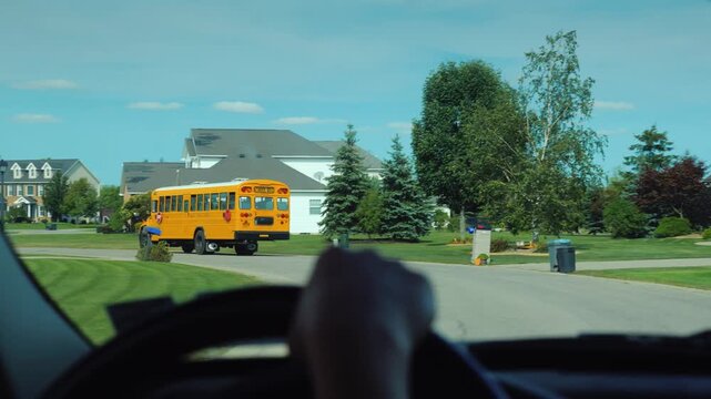 First person view driving car through quiet suburban residential neighborhood street following yellow school bus transporting children to educational facility during bright sunny day on summer morning